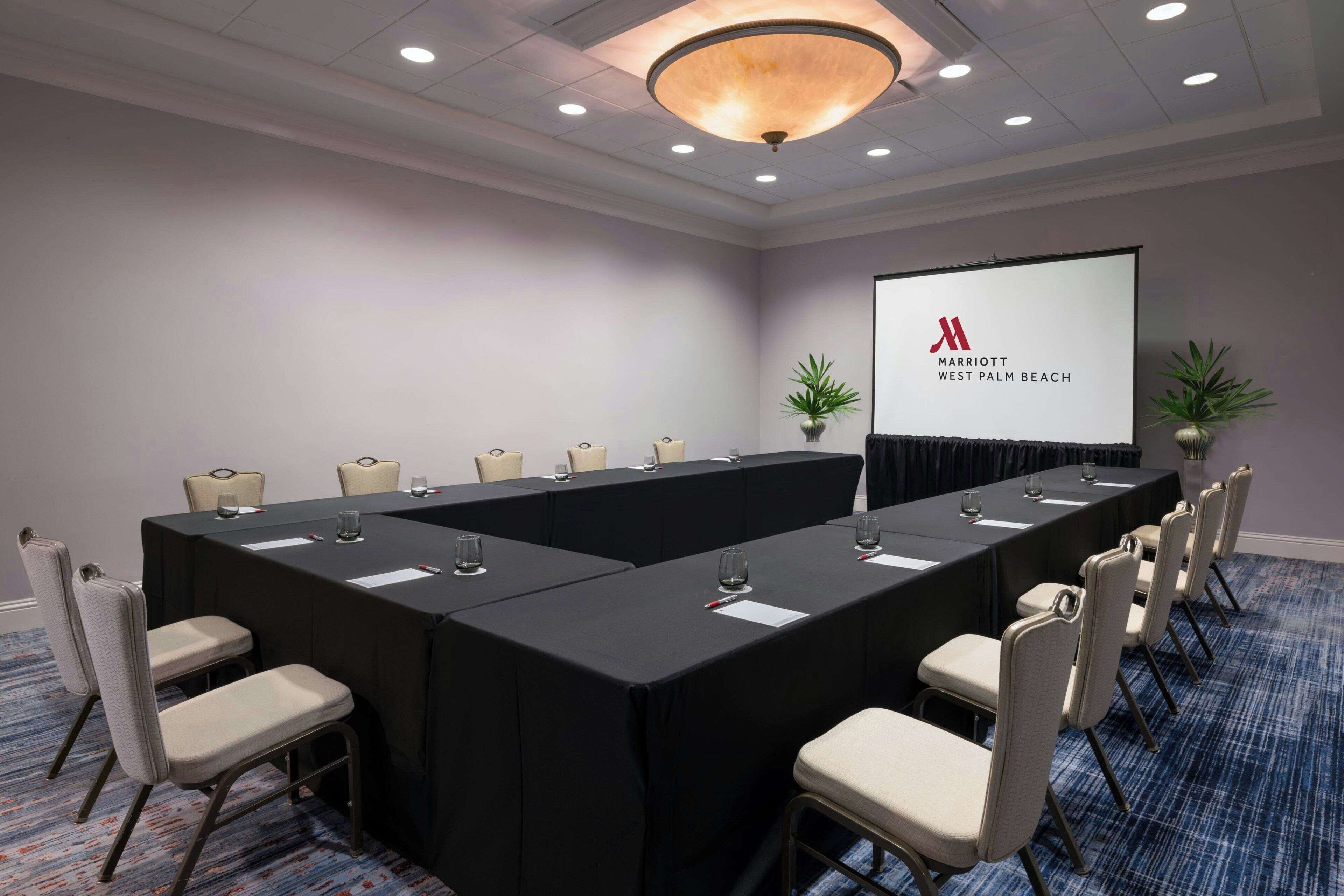 Een congreszaal met een lange tafel, stoelen, een projectiescherm en het logo van Marriott West Palm Beach.