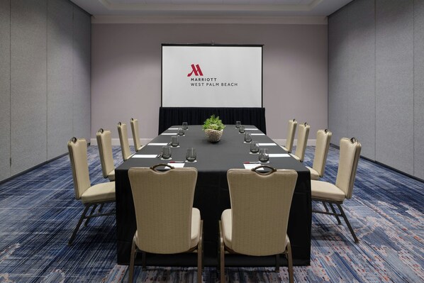 Un salón de conferencias con una larga mesa, sillas color beige, una pantalla de proyector y un logotipo en la pantalla.