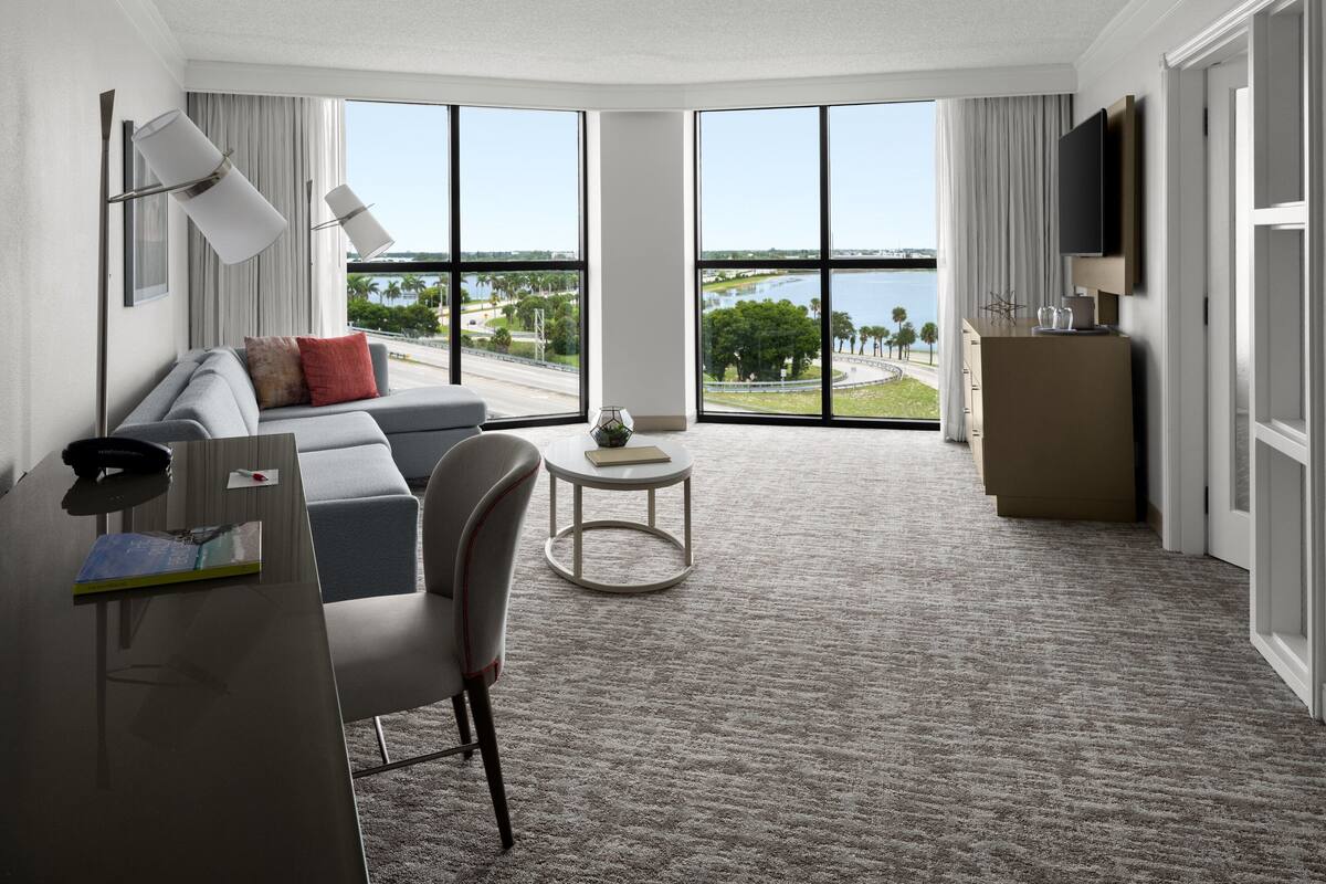 Een moderne hotelkamer met een groot raam met uitzicht op een brug en groen, een bank, een bureau met stoel, een klein rond tafeltje en een aan de muur bevestigde televisie.