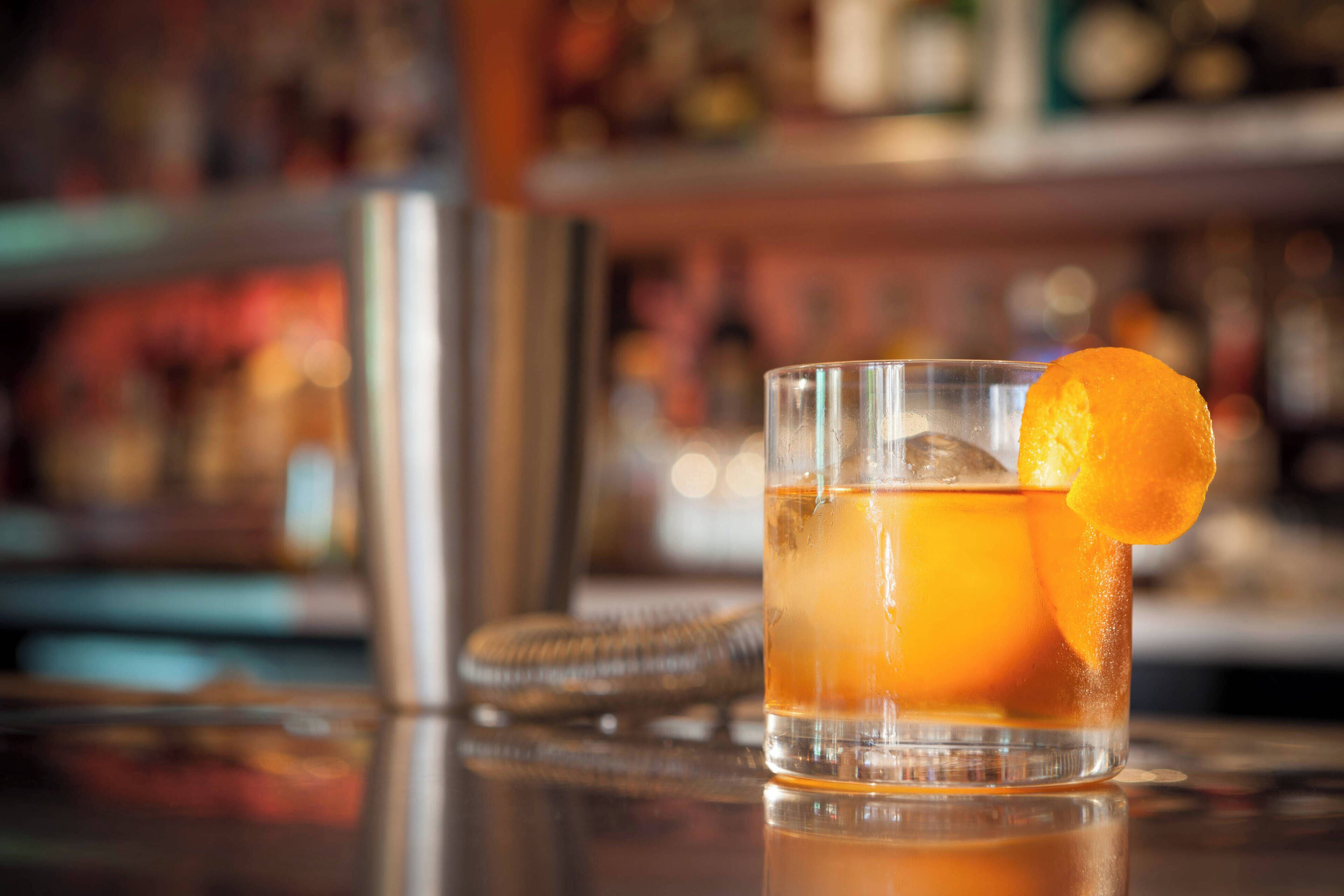 Un verre contenant une boisson de couleur ambrée, garnie d’une tranche d’orange, un shaker et, en arrière-plan, une étagère floue remplie de différentes bouteilles.