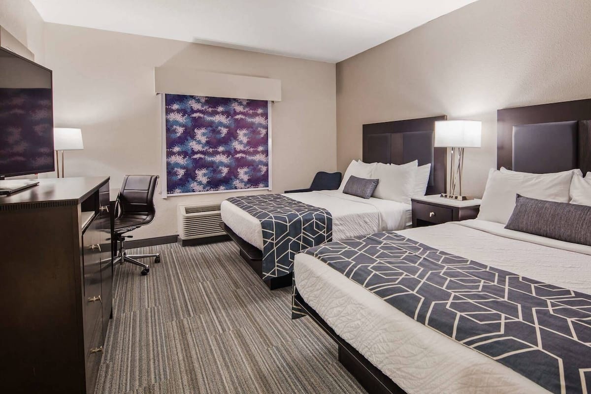 A hotel room with two beds, a desk, a chair, a large abstract painting, and a window.