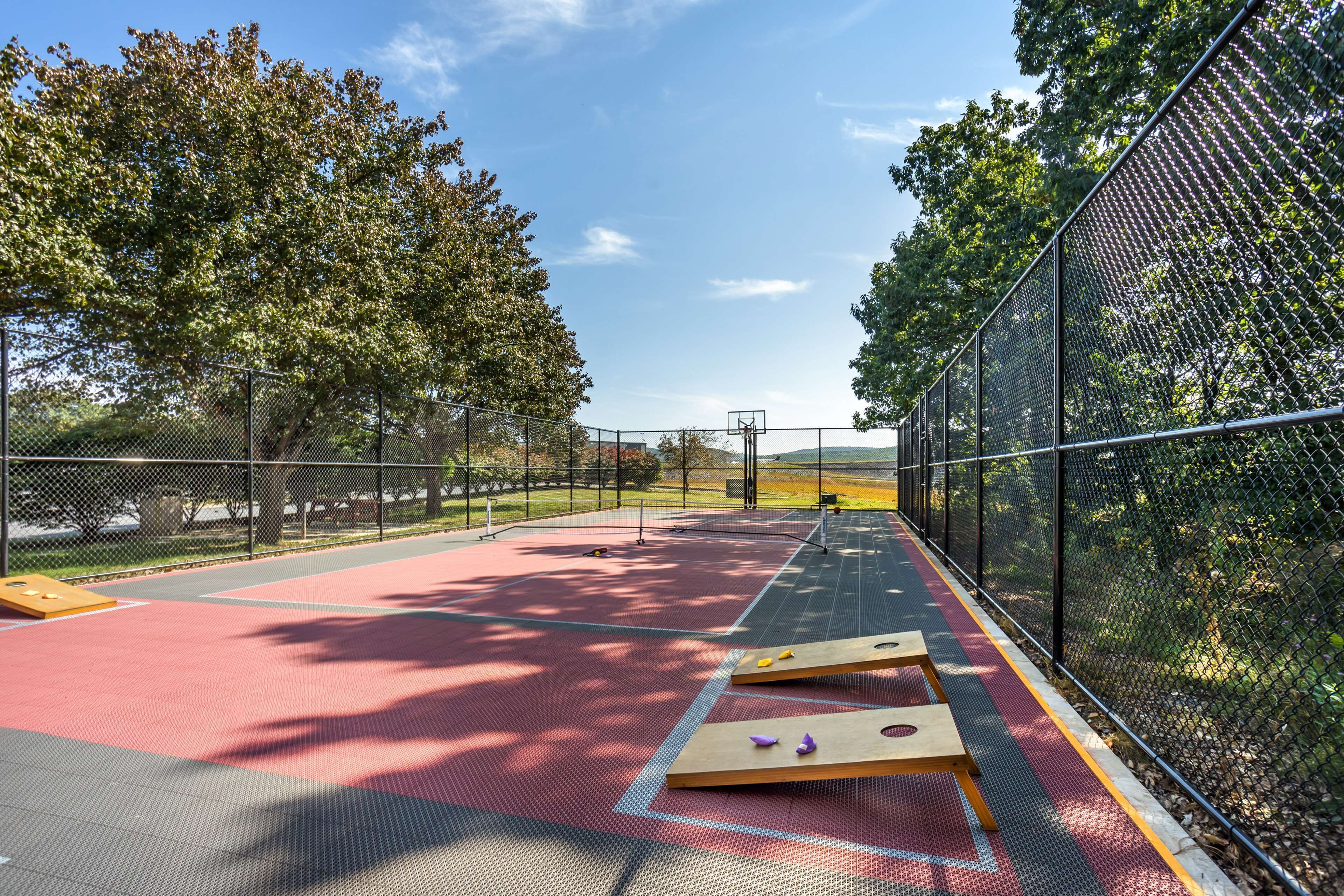 Quadra de basquete ao ar livre com superfície vermelha, cercada por uma grade e árvores.