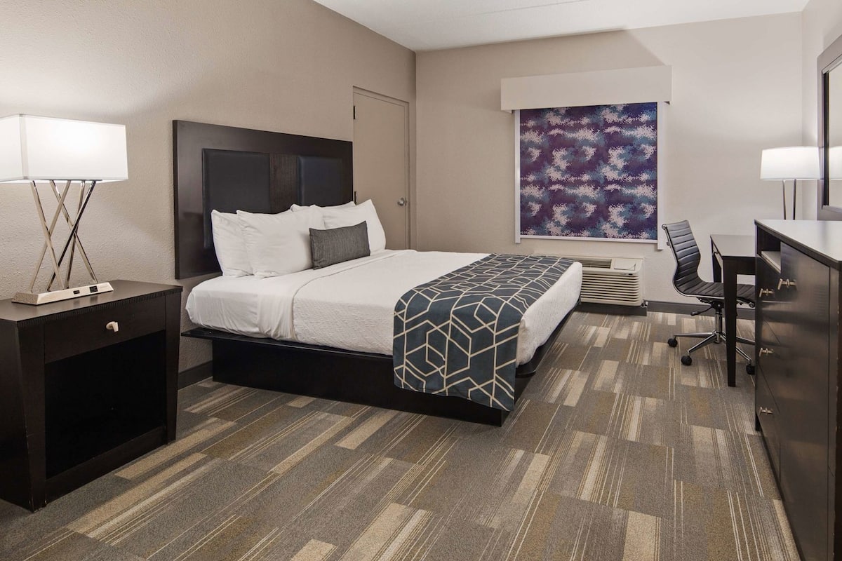 A hotel room with a bed, desk, chair, TV, and artwork on the wall.