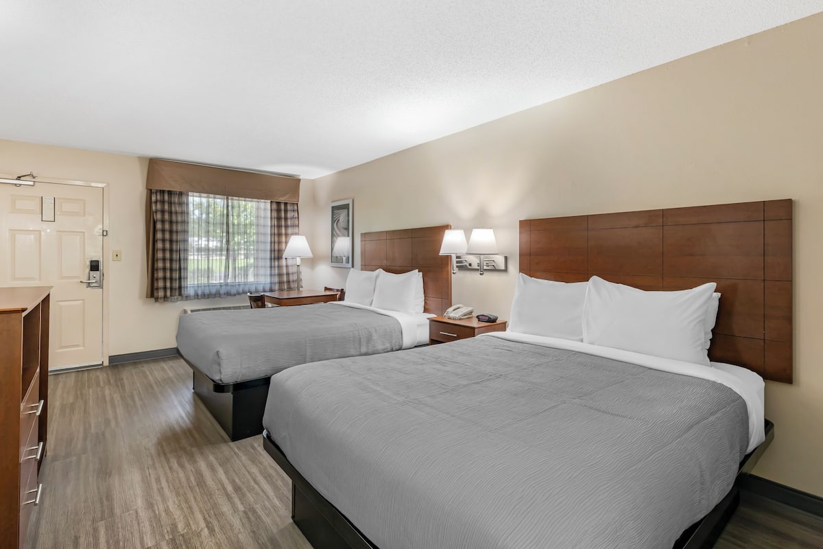 A hotel room with two beds, a desk, a chair, a window with curtains, and a wooden headboard.