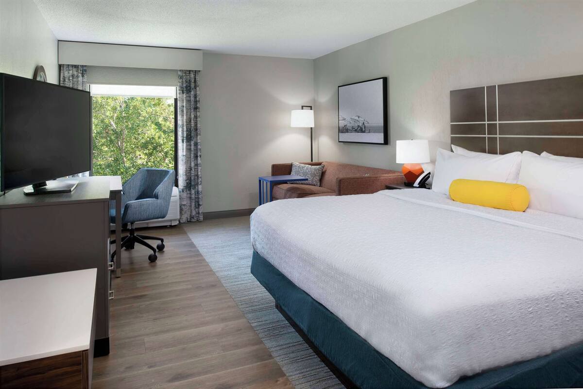 Une chambre d’hôtel comprenant un lit, un bureau avec une télévision, une chaise, un canapé et une fenêtre avec des rideaux.