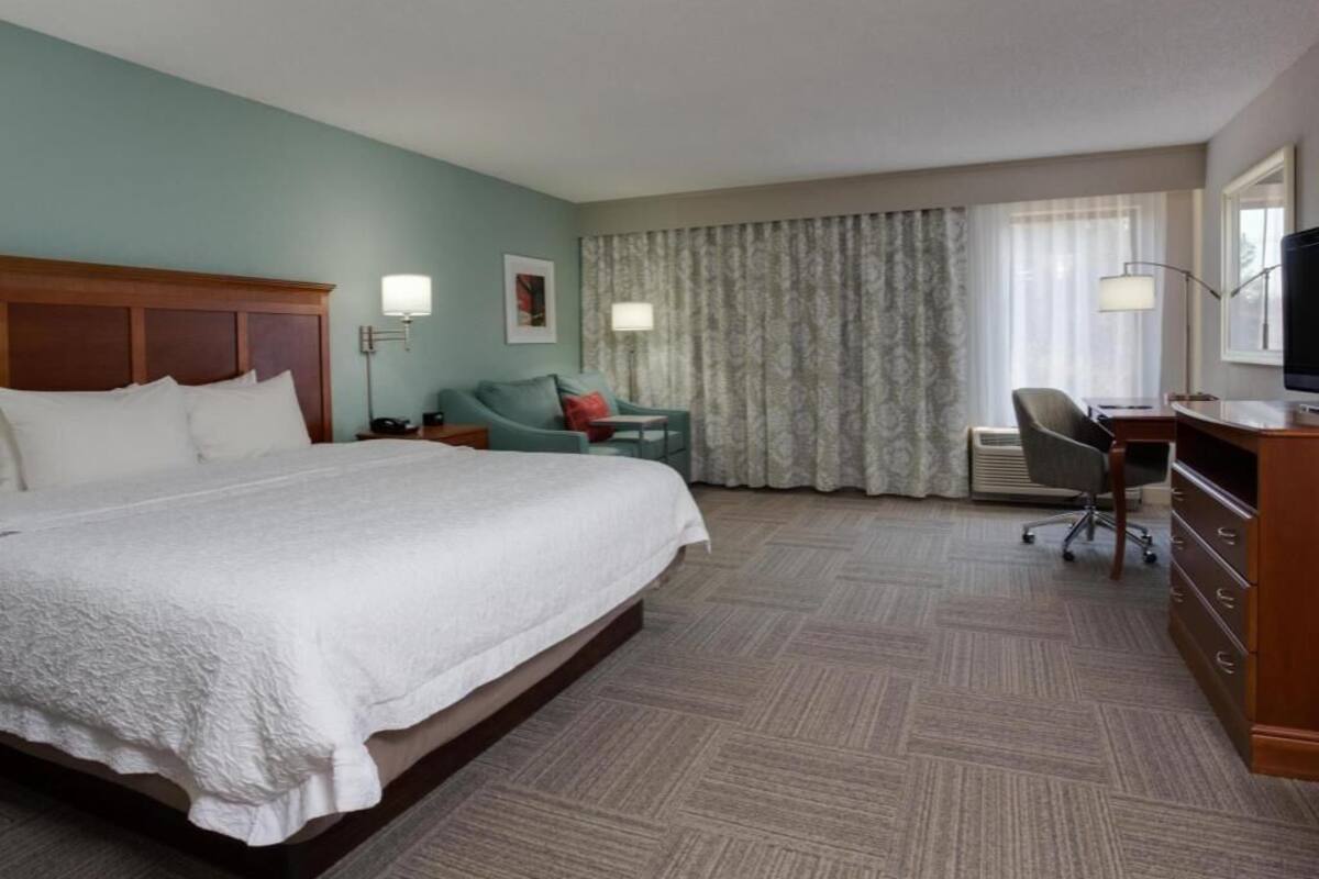 Une chambre d’hôtel avec un grand lit, un bureau, une chaise, une lampe et une fenêtre avec des rideaux.
