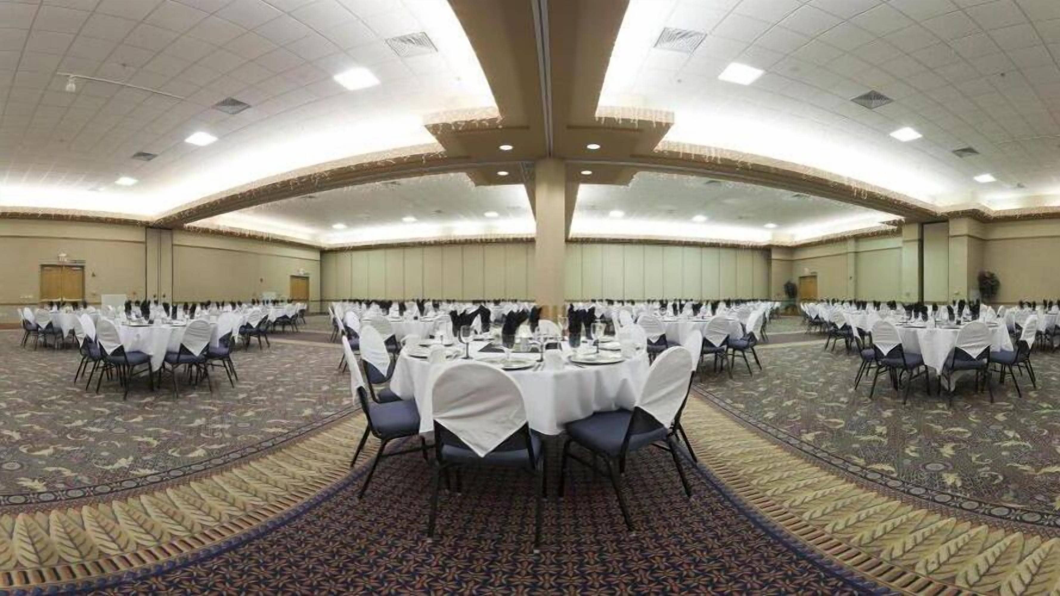 A large banquet hall with round tables covered in white tablecloths, arranged in rows.