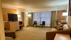 A hotel room with a flat-screen TV, a wooden cabinet, a sofa, a dining table with chairs, and a large window with curtains. - Holiday Inn Spearfish - Convention Center by IHG (Spearfish)