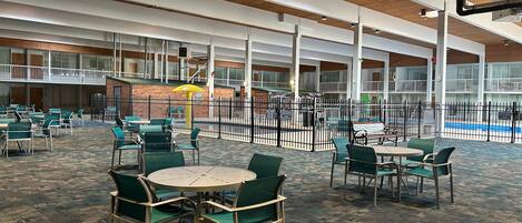 A spacious indoor area with multiple tables and chairs, a pool in the background, and a high ceiling with wooden beams.