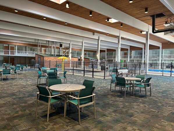 A spacious indoor area with multiple tables and chairs, a pool in the background, and a high ceiling with wooden beams.