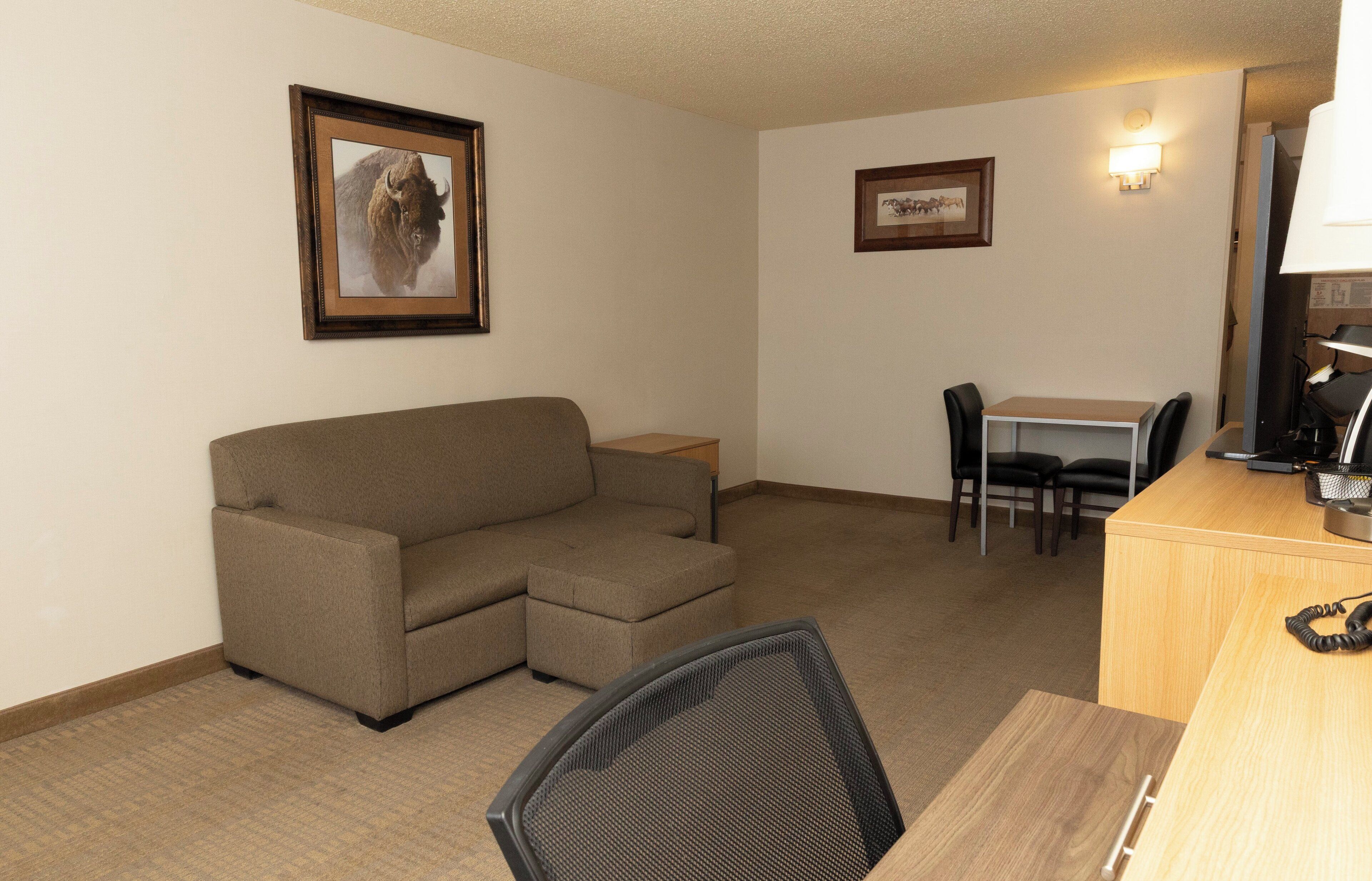 Een gemeubileerde kamer met een bank, een eettafel met stoelen, een bureau met een computer, en lijstjes met afbeeldingen aan de muur.