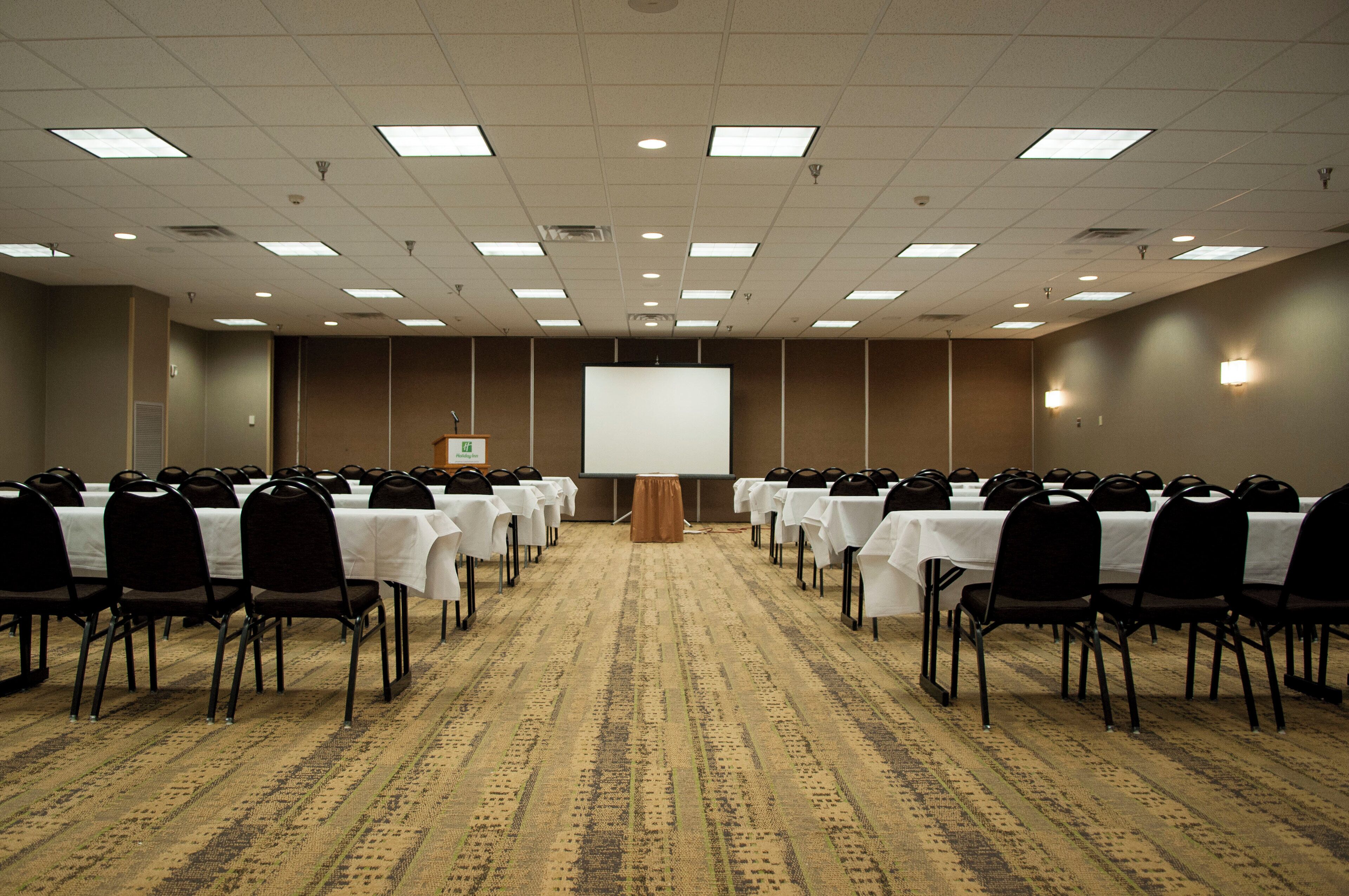 A conference room with rows of tables and chairs, a podium, and a projection screen.