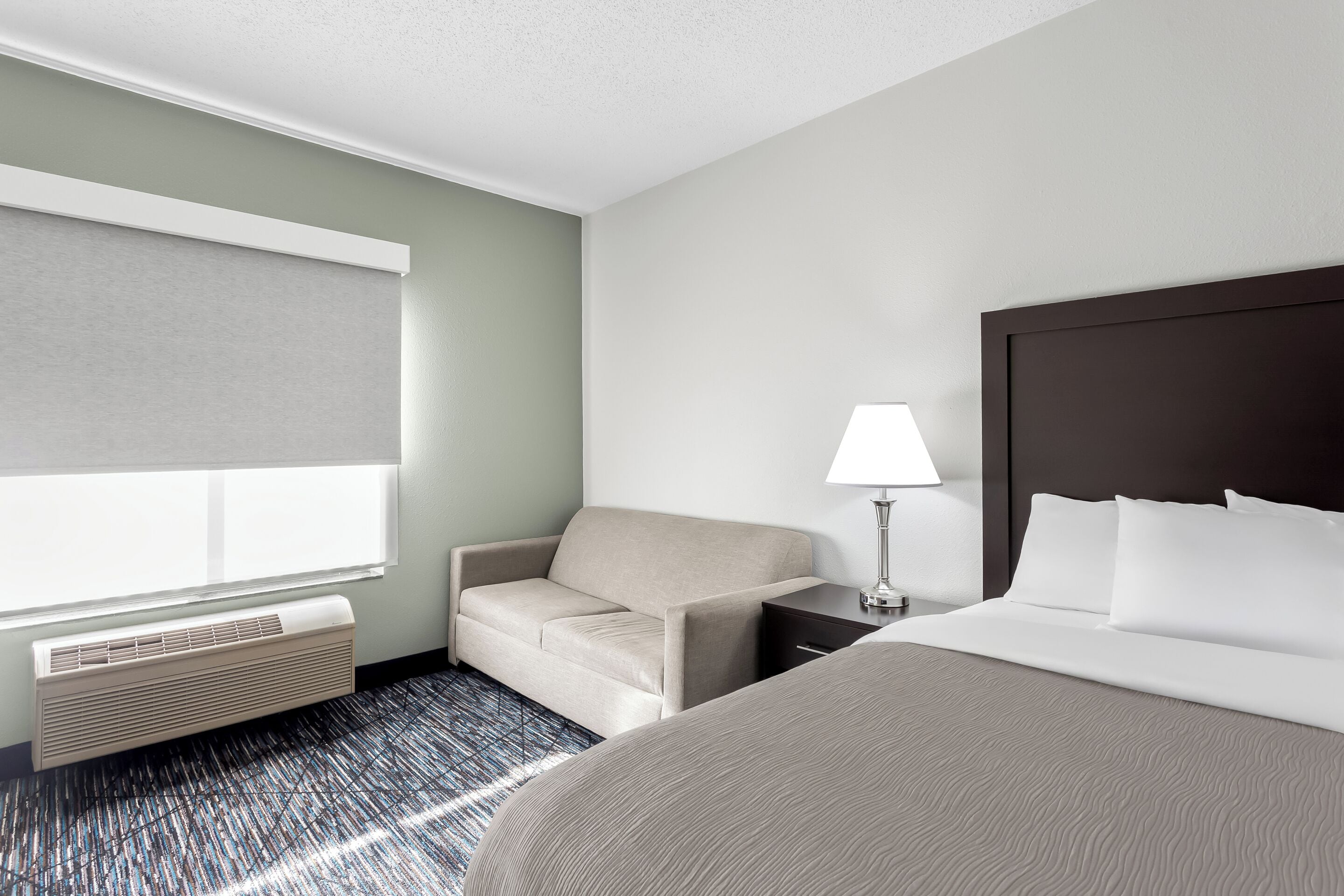 Een hotelkamer met een bed, een sofa, een nachtkastje, een lamp en een raam met jaloezieën.