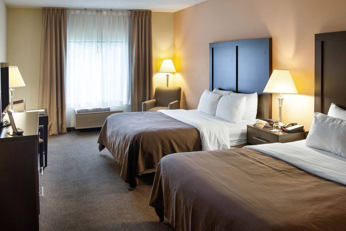 Habitación de hotel con dos camas, un escritorio, una silla, lámparas y un ventanal con cortinas.