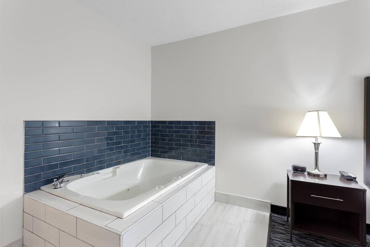 Un baño moderno con una bañera independiente, una mesita de noche de madera oscura con una lámpara, y un piso de baldosas blancas.