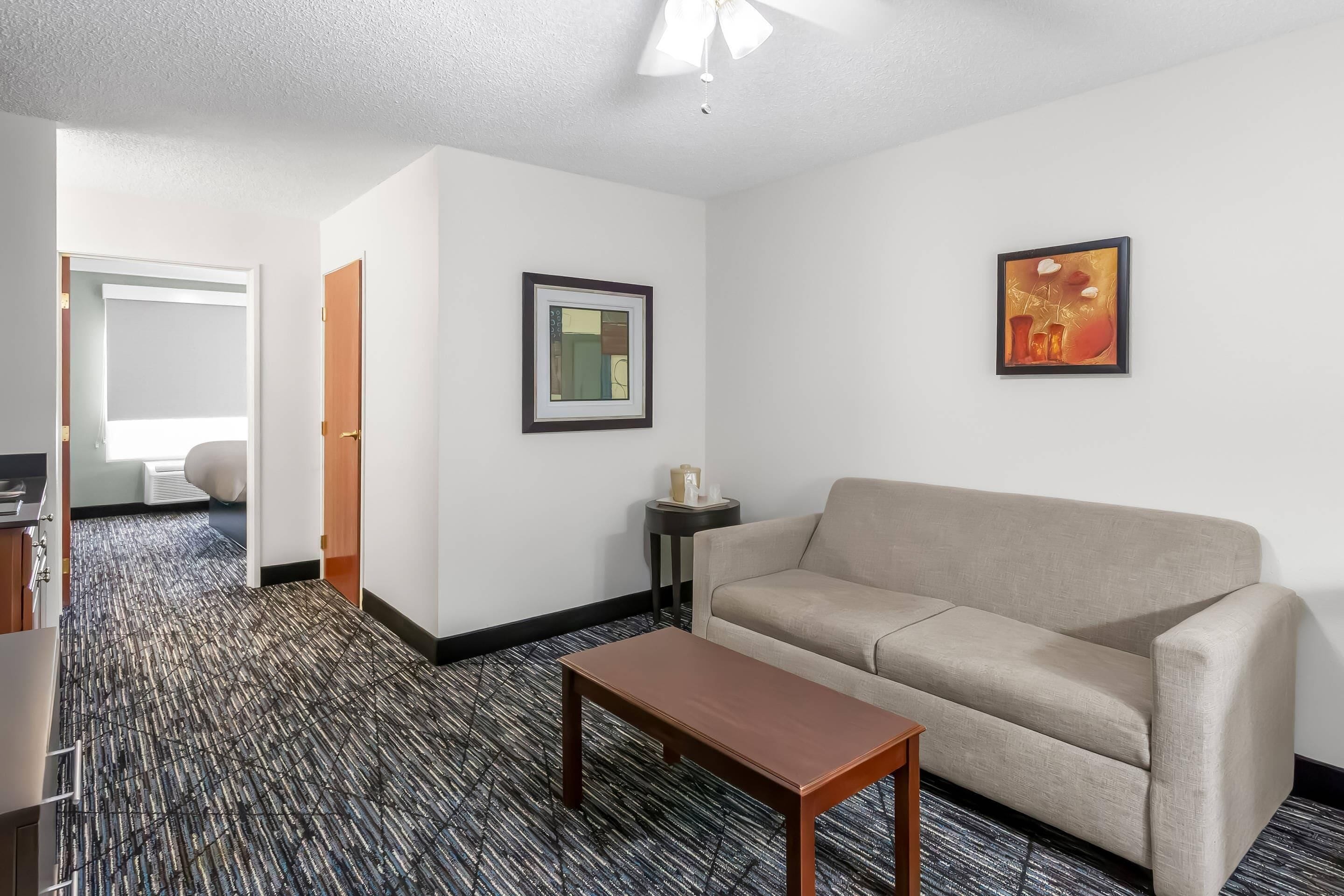 Habitación de hotel con un sofá color beige, una mesa de centro de madera, un cuadro enmarcado en la pared y piso cubierto de alfombra.