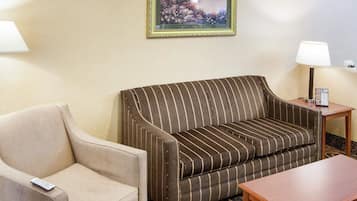 Una habitación de hotel con un sofá a rayas, un sillón sencillo, una mesita de madera con una lámpara y un cuadro en la pared.