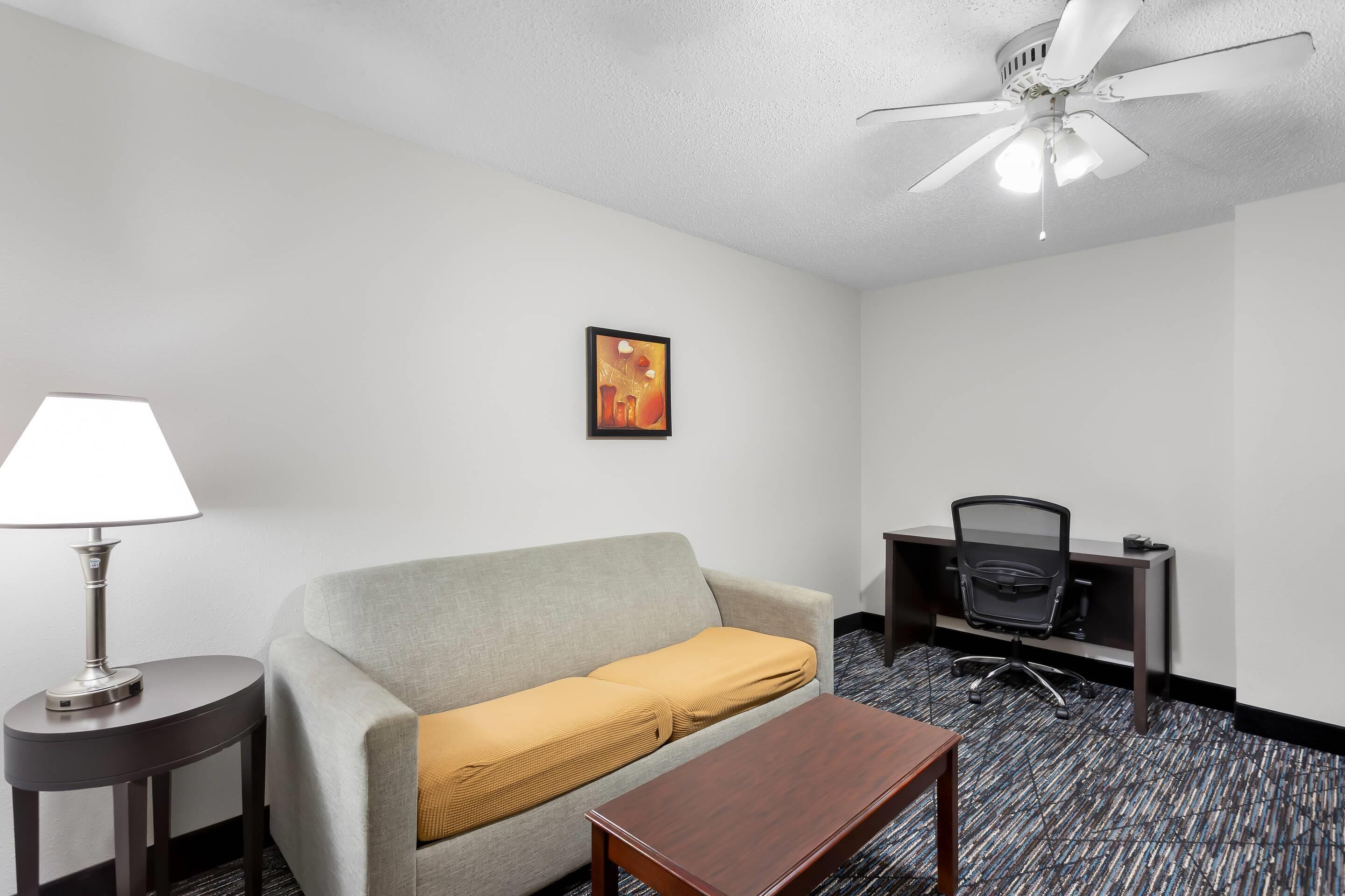 A hotel room with a gray sofa, a wooden coffee table, a desk with a chair, a lamp, and a ceiling fan.