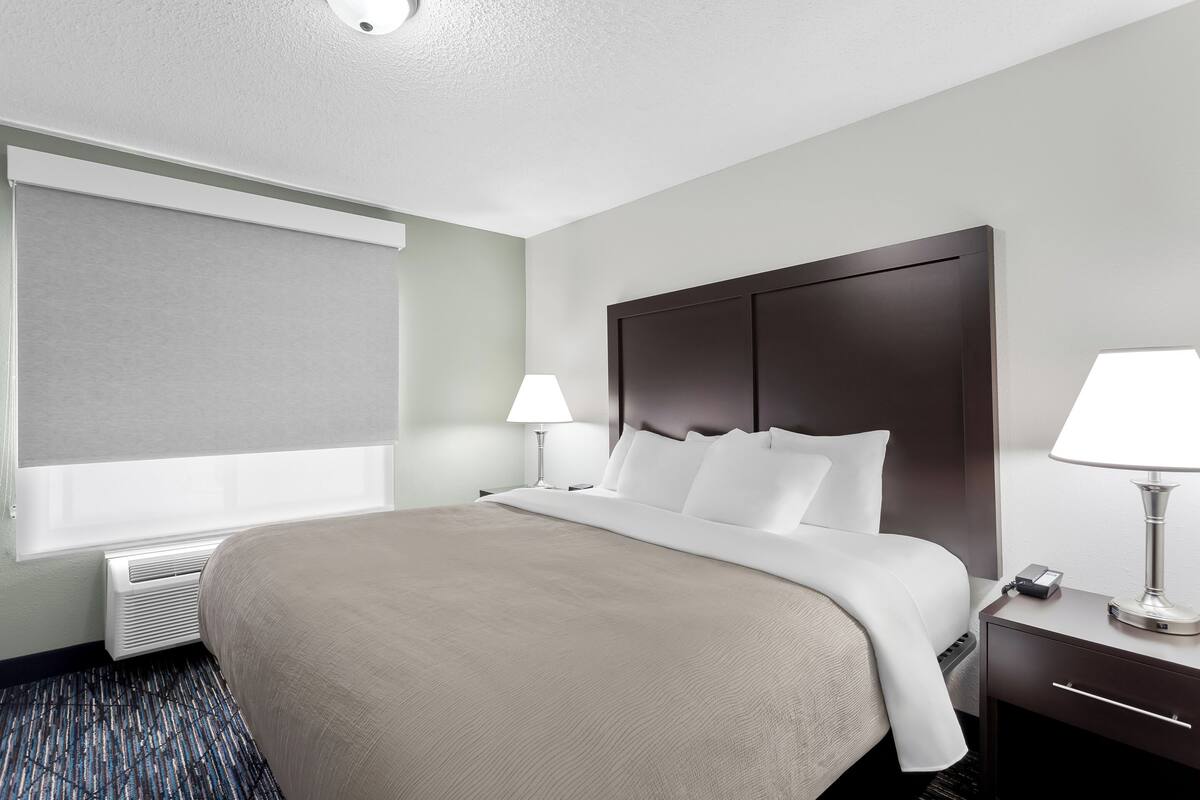 Habitación de hotel con una cama grande, una mesita de noche con lámpara, una ventana con persiana y un calentador de pared.