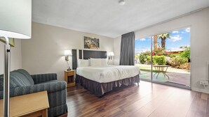 A hotel room with a bed, a chair, a nightstand, a lamp, and a view of a garden through a sliding glass door.