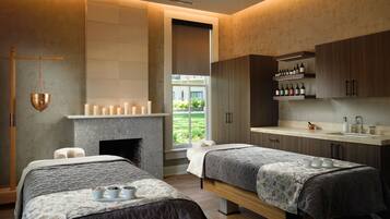 A modern bedroom with two beds, a fireplace, a large window, and a bathroom with a sink and shelves.