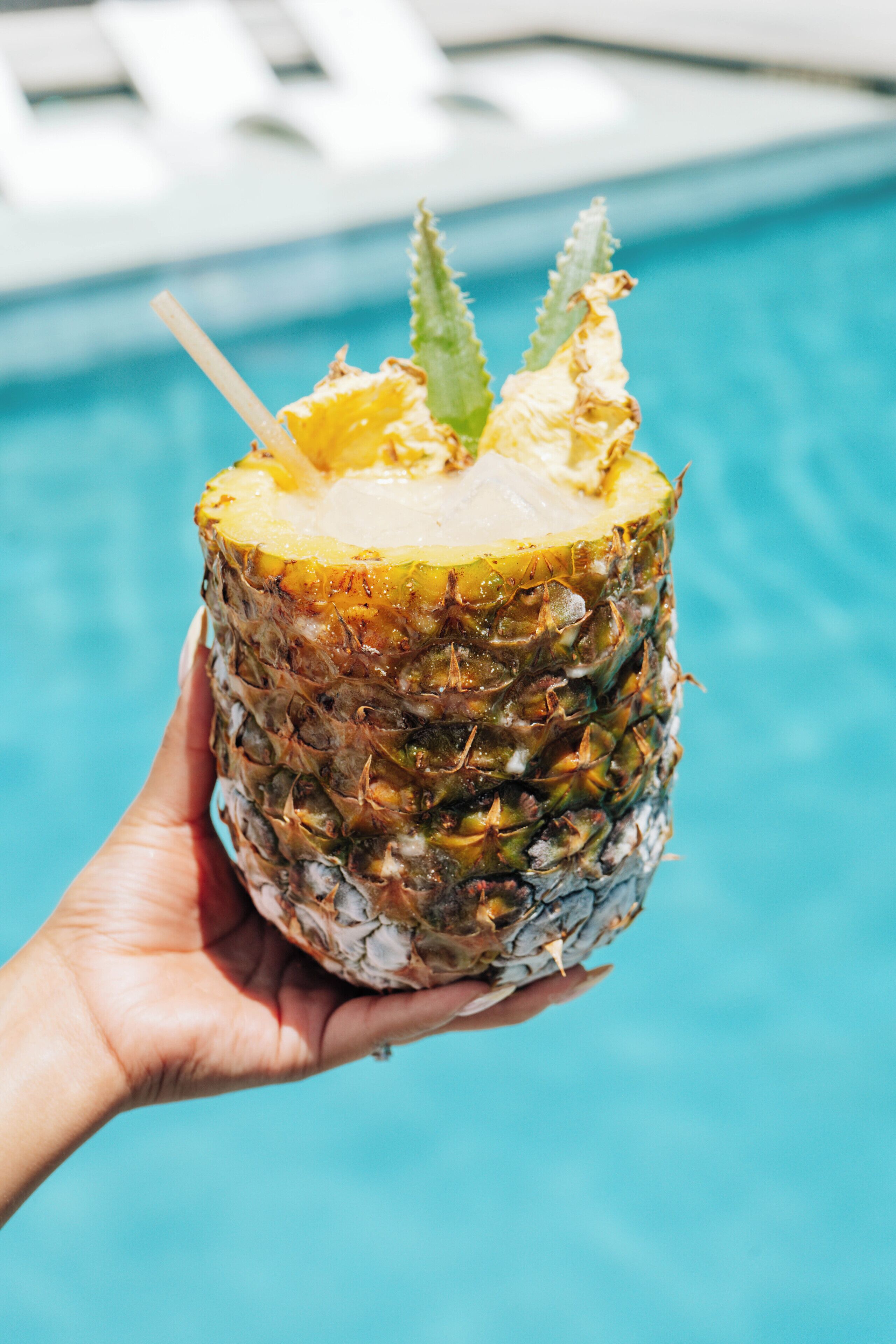 Eine Hand hält eine Ananas mit einem Strohhalm, im Hintergrund ist ein Schwimmbecken zu sehen.