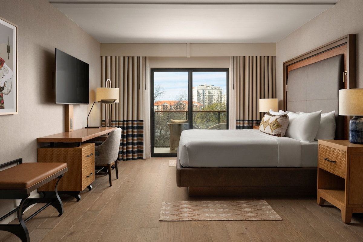 A hotel room with a bed, desk, chair, TV, and a balcony with a view of buildings.
