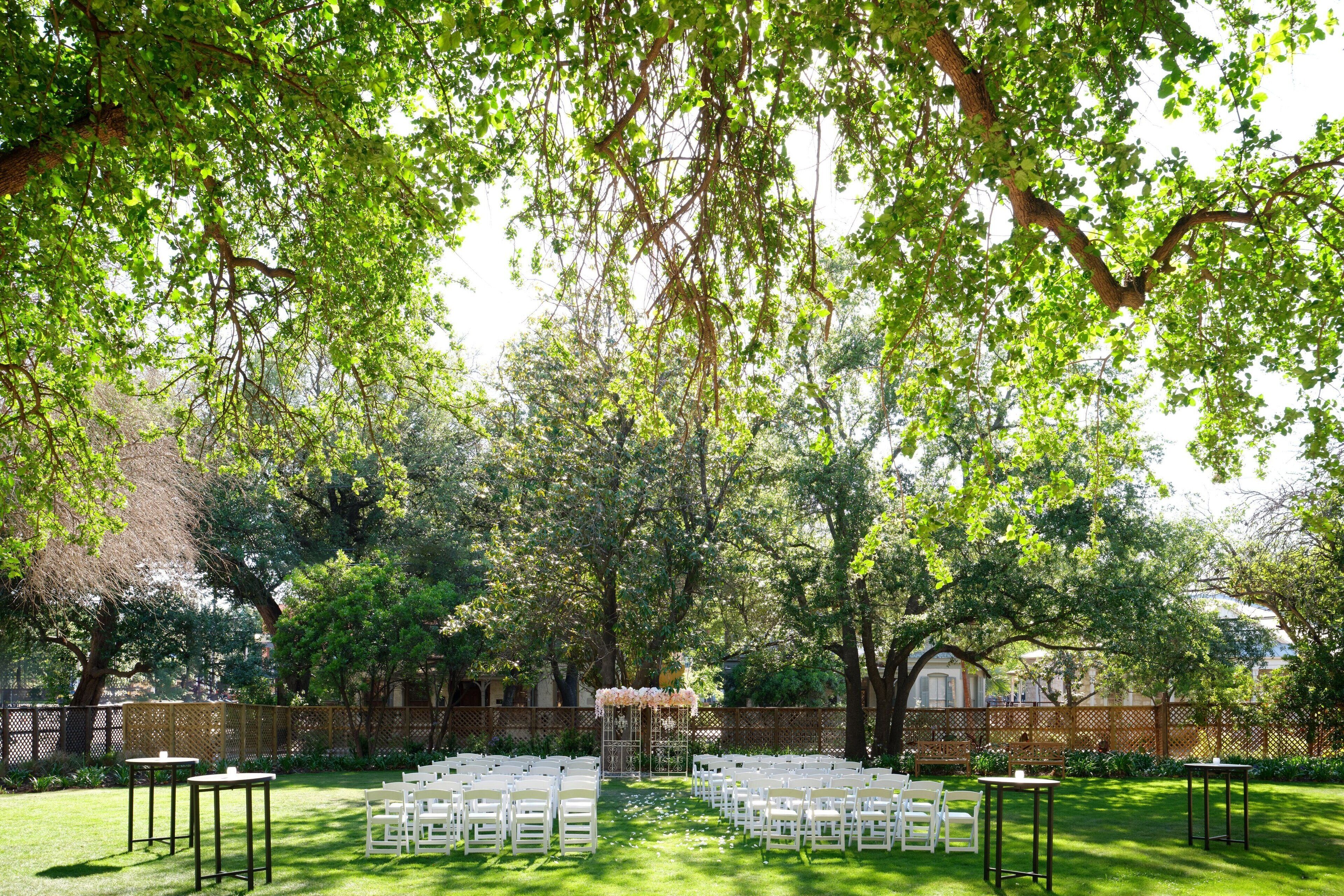 An outdoor wedding venue with rows of white chairs and tables, surrounded by lush greenery and trees.
