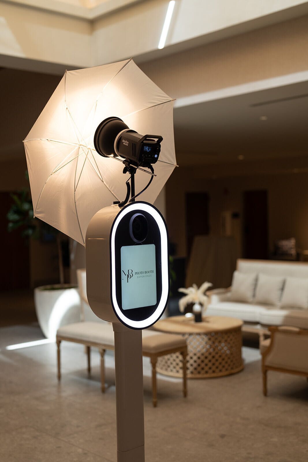 A modern photo booth with a light umbrella and a camera, set in a well-lit living area with a sofa and decorative items.