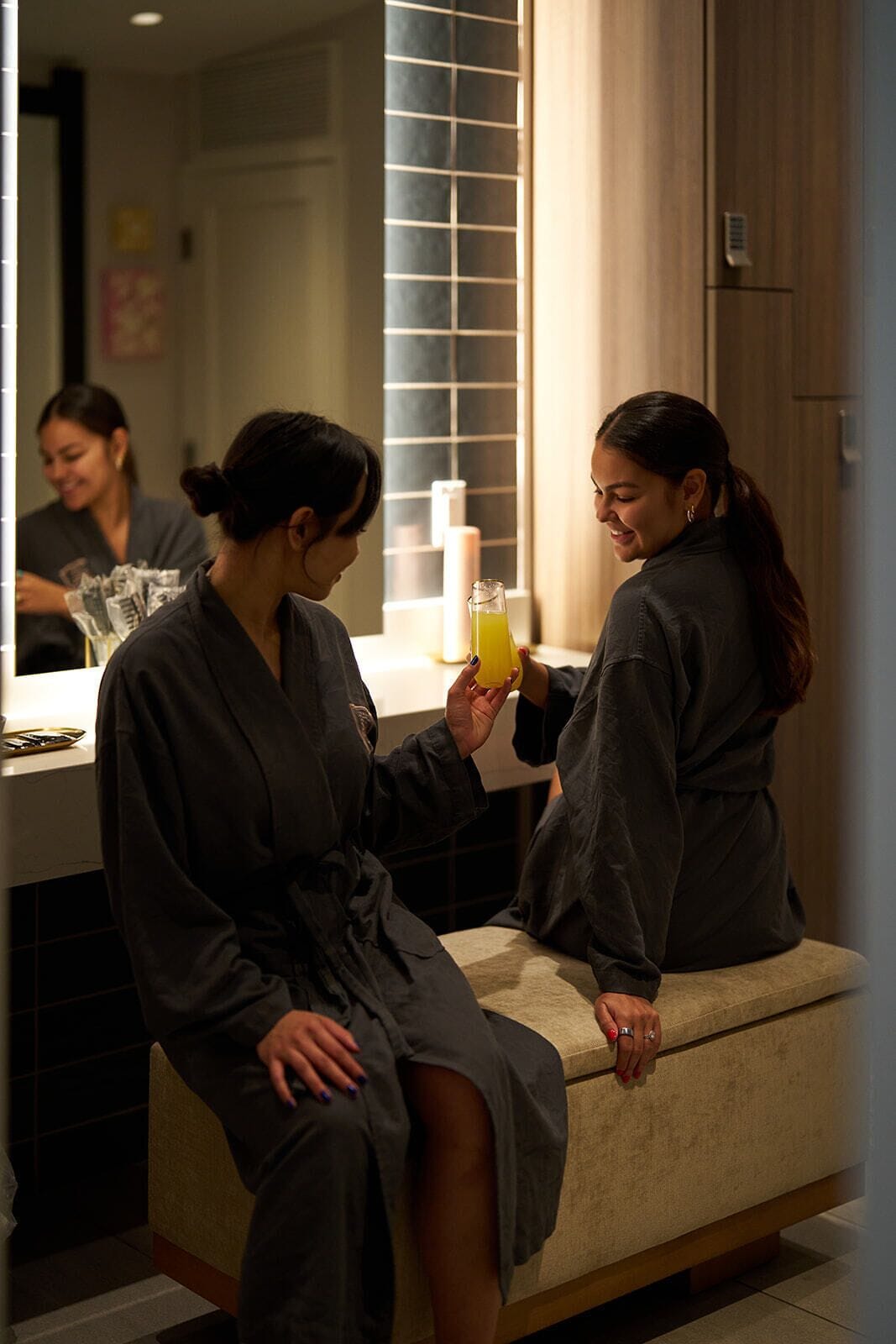 Two individuals in a room with a bench, a mirror, and a window with blinds.