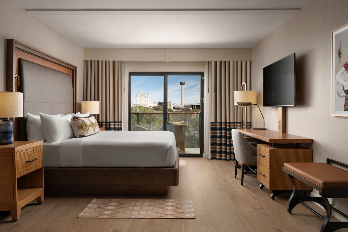 A hotel room with a bed, desk, chair, TV, and a view of a tower through the window.