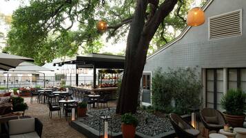 Un patio al aire libre con mobiliario, un árbol y un restaurante al fondo.