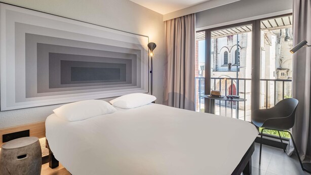 A modern hotel room with a large bed, a small table with a chair, a view of the city through large windows, and a unique wall art piece.
