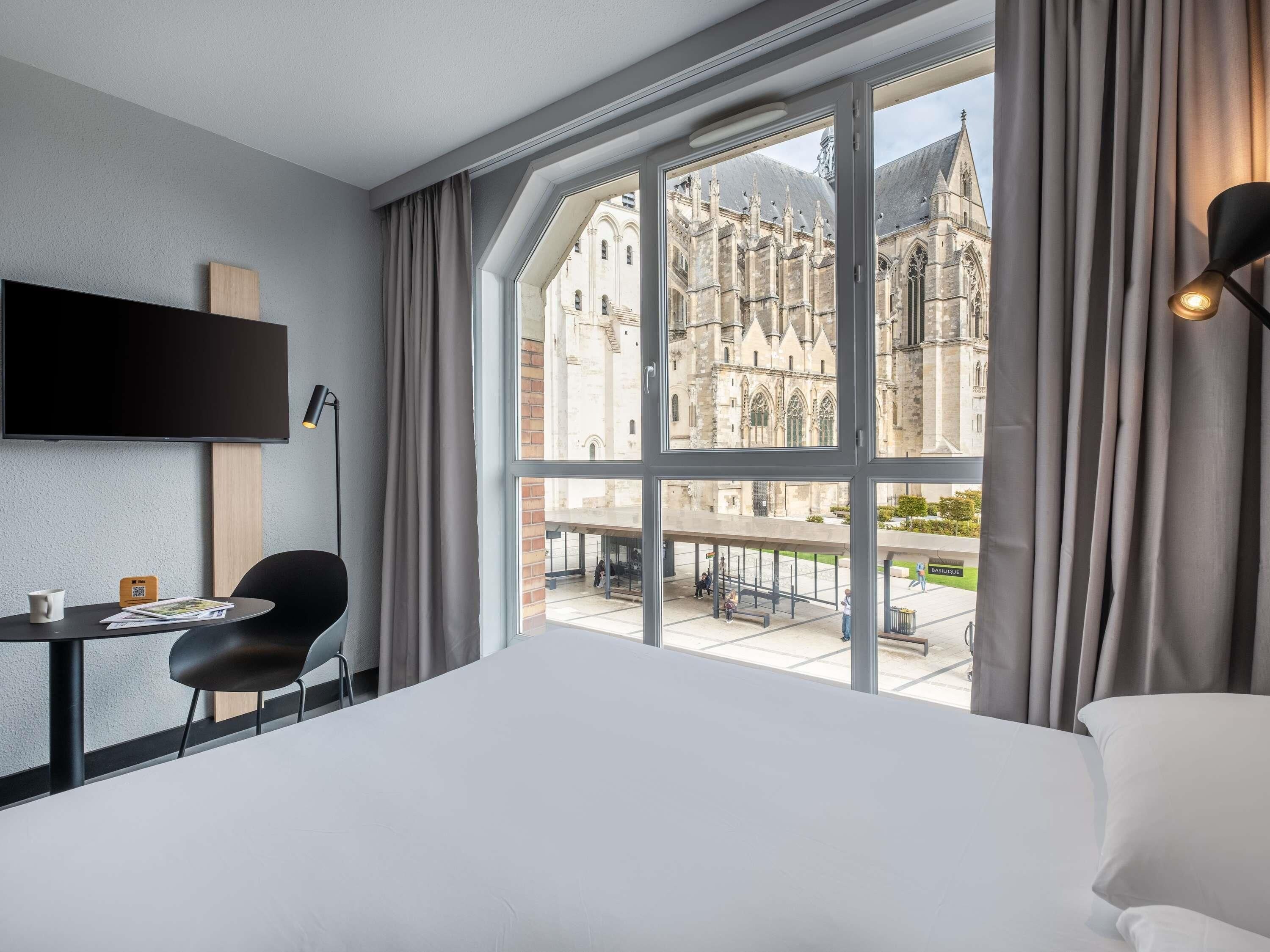 A hotel room with a large window, a bed, a desk with a chair, a flat-screen TV, and a coffee table.
