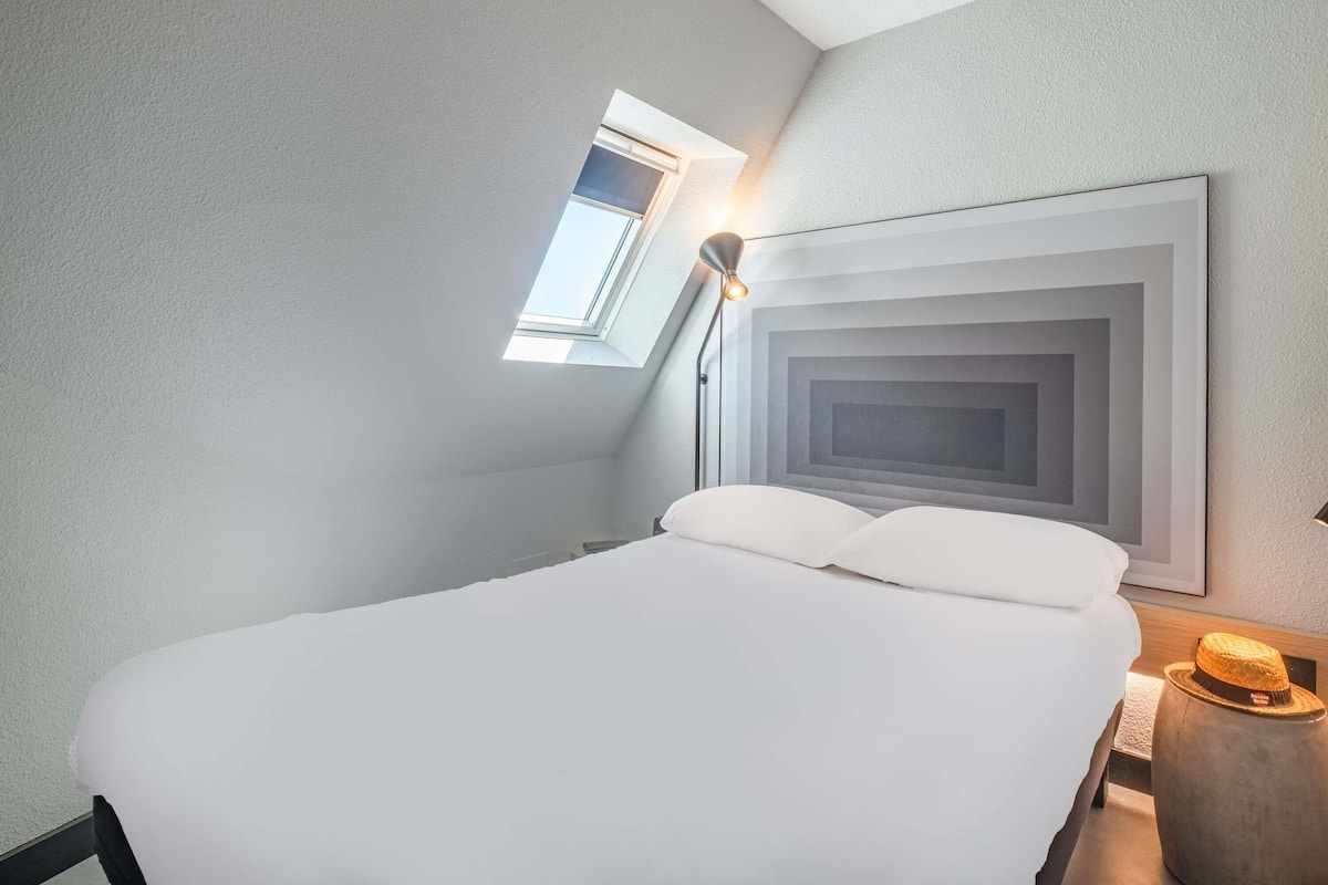 Quarto de hotel moderno com claraboia, uma cama grande e uma obra de arte minimalista na parede.