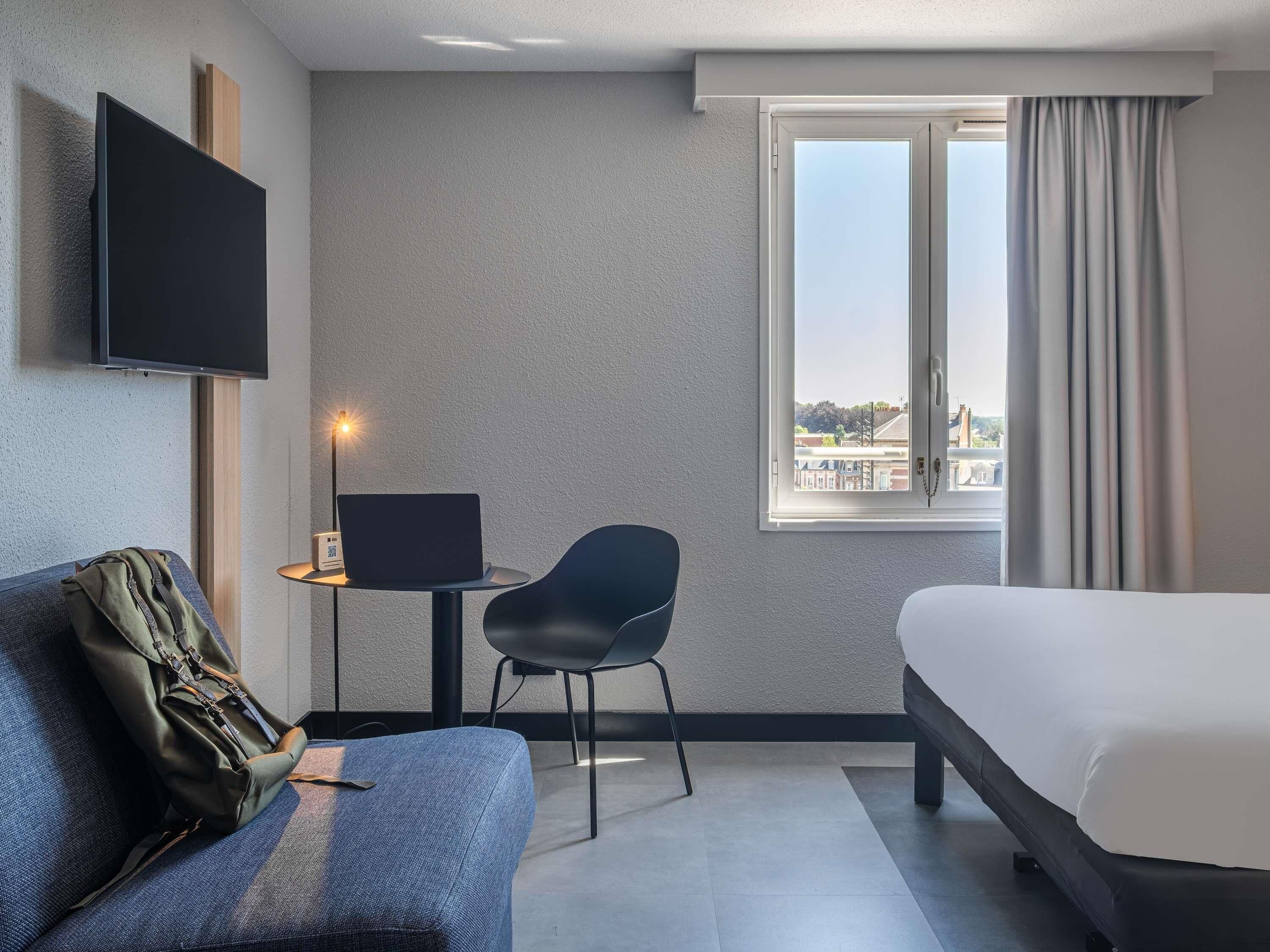Ein Hotelzimmer mit einem Bett, einem Schreibtisch mit Laptop, einem Stuhl, einem an der Wand befestigten Fernseher und einem Fenster mit Vorhängen.