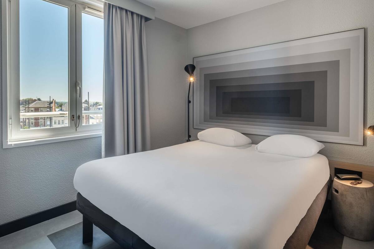 A hotel room with a large bed, a view of the city through a window, and a modern wall art piece.