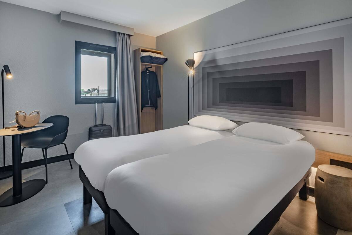 A modern hotel room with a large bed, a desk, a chair, a wardrobe, and a bedside lamp.