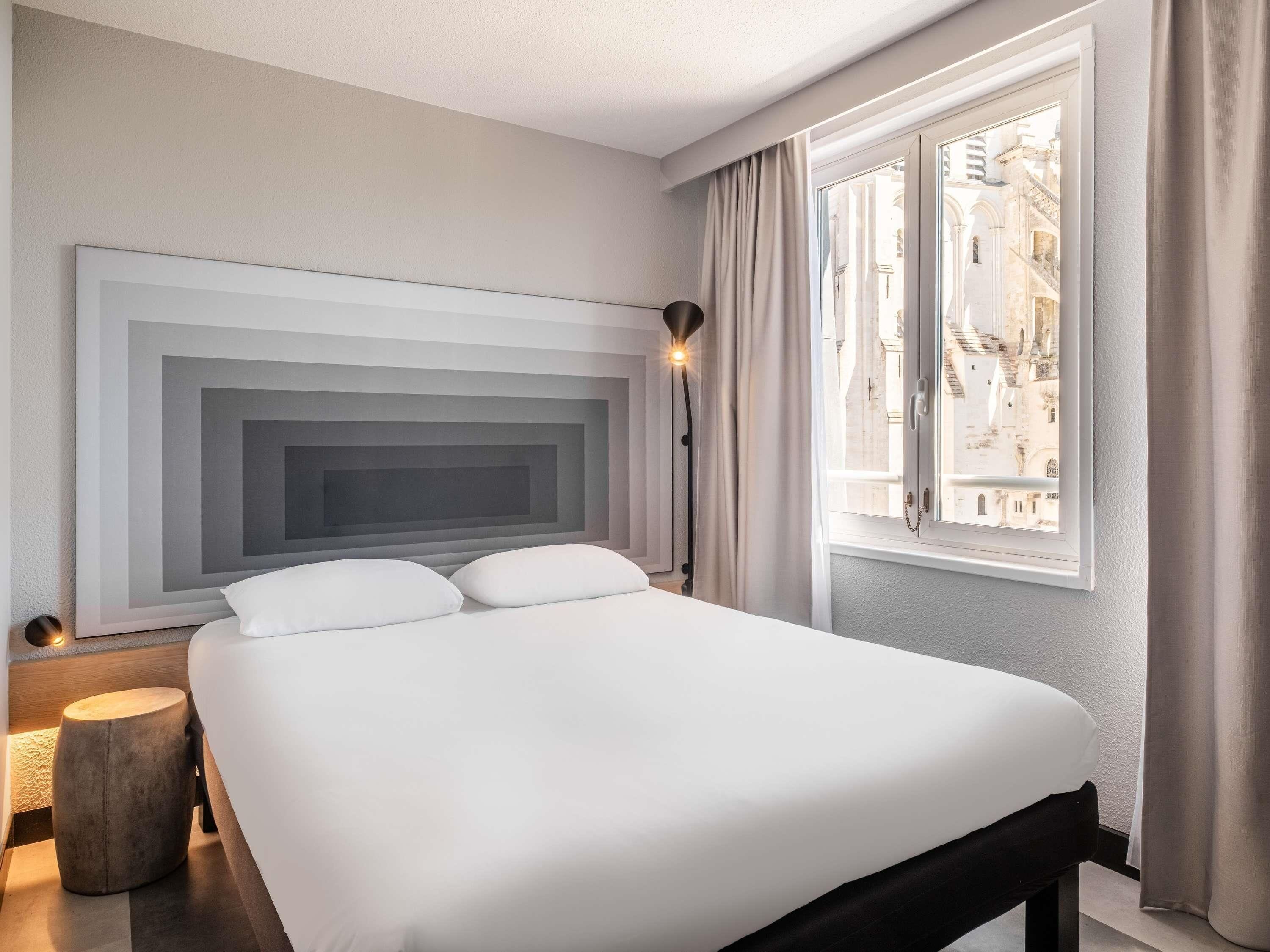 A modern hotel room with a large bed, a headboard with a geometric design, a bedside lamp, and a window with a view of buildings.
