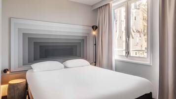 A modern hotel room with a large bed, a headboard with a geometric design, a bedside lamp, and a window with a view of buildings.