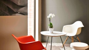A modern room with a brick wall, a large window, a white table with a potted orchid, and two chairs (one orange, one white).