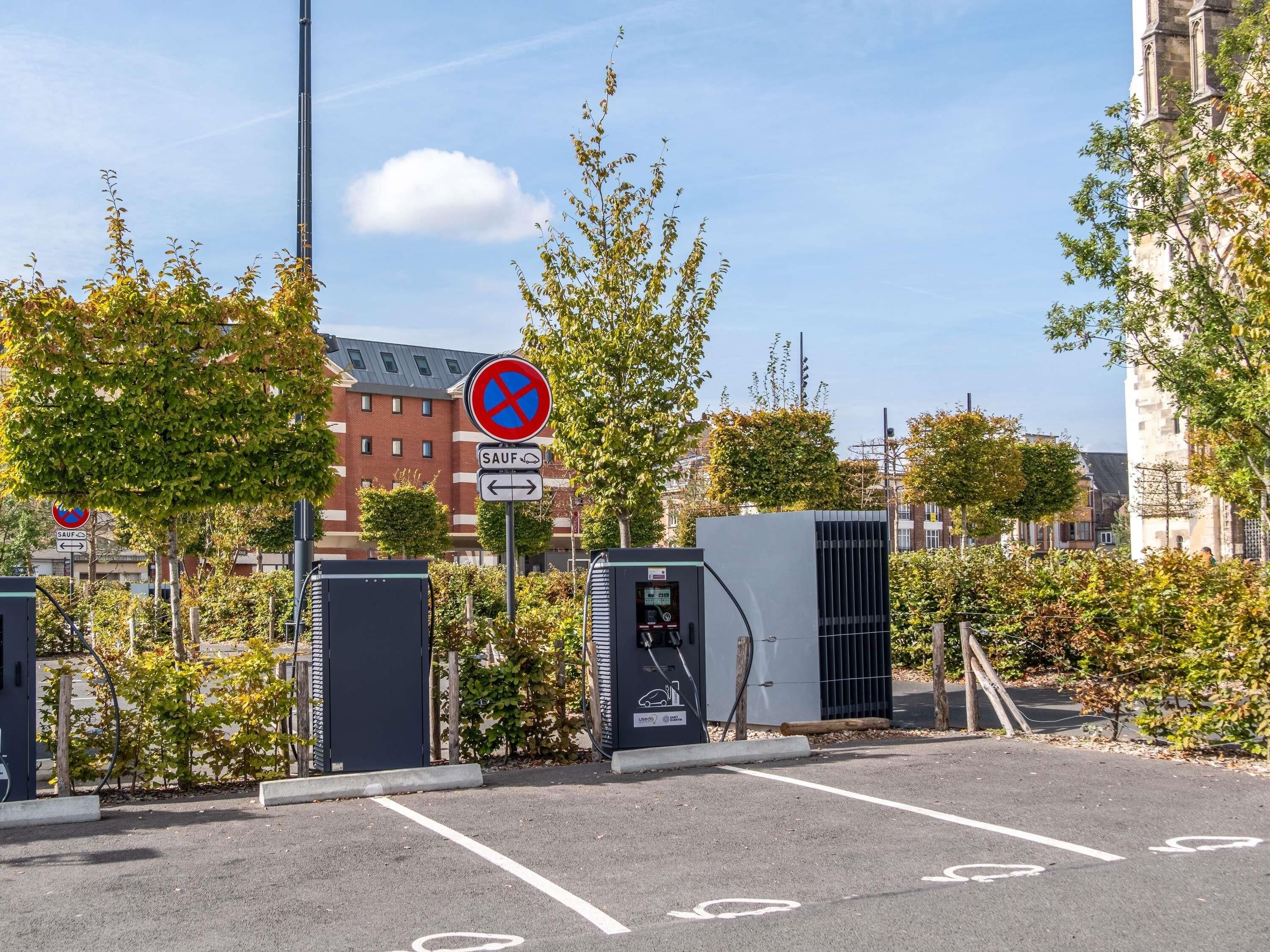 A parking area with a no parking sign, electric vehicle charging station, and a building in the background.
