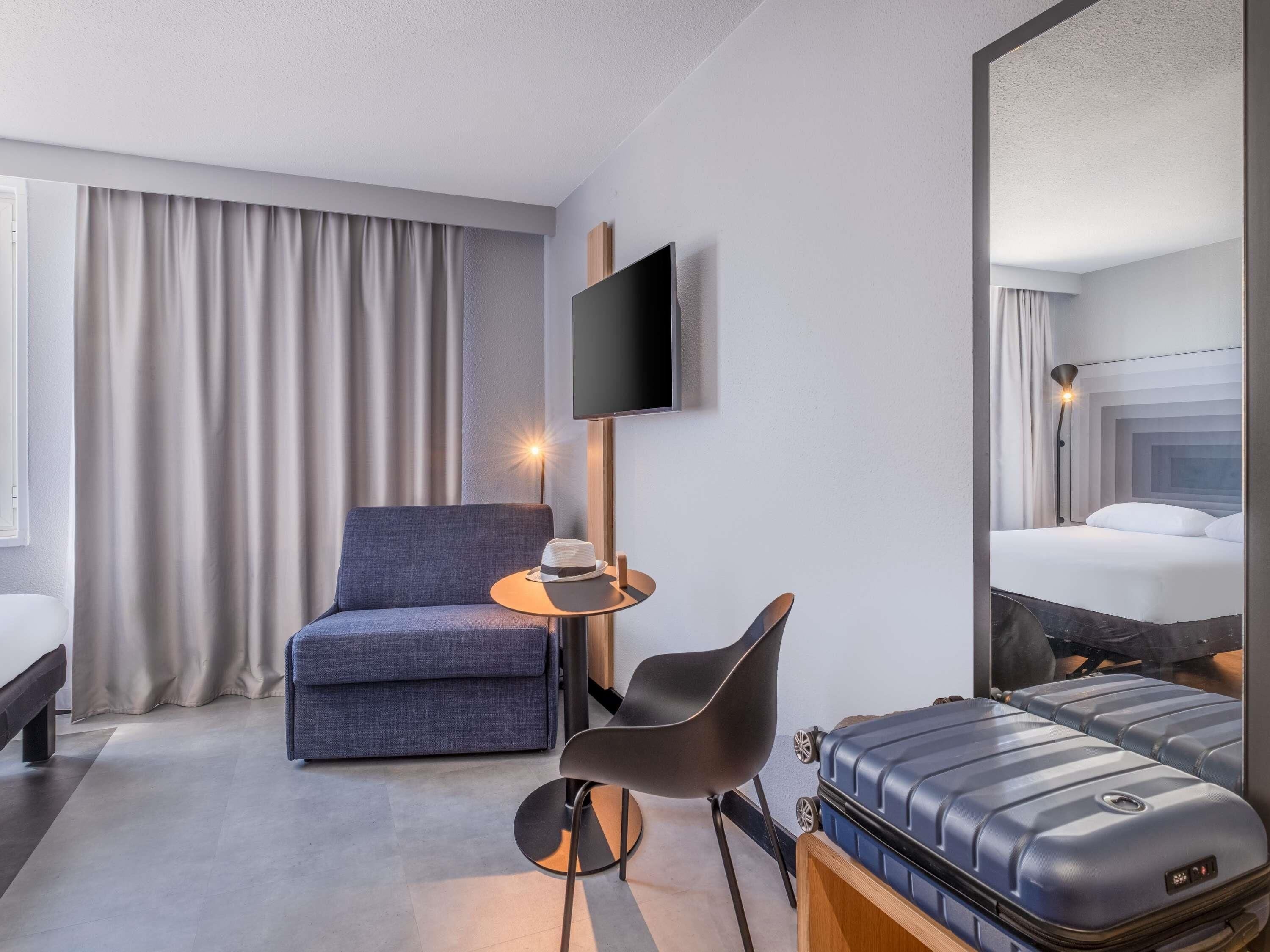 Et hotelværelse med en seng, en stol, et lille bord med en hat, et fjernsyn ophængt på væggen og en kuffert.