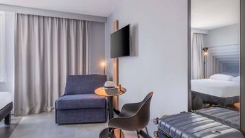 Et hotelværelse med en seng, en stol, et lille bord med en hat, et fjernsyn ophængt på væggen og en kuffert.