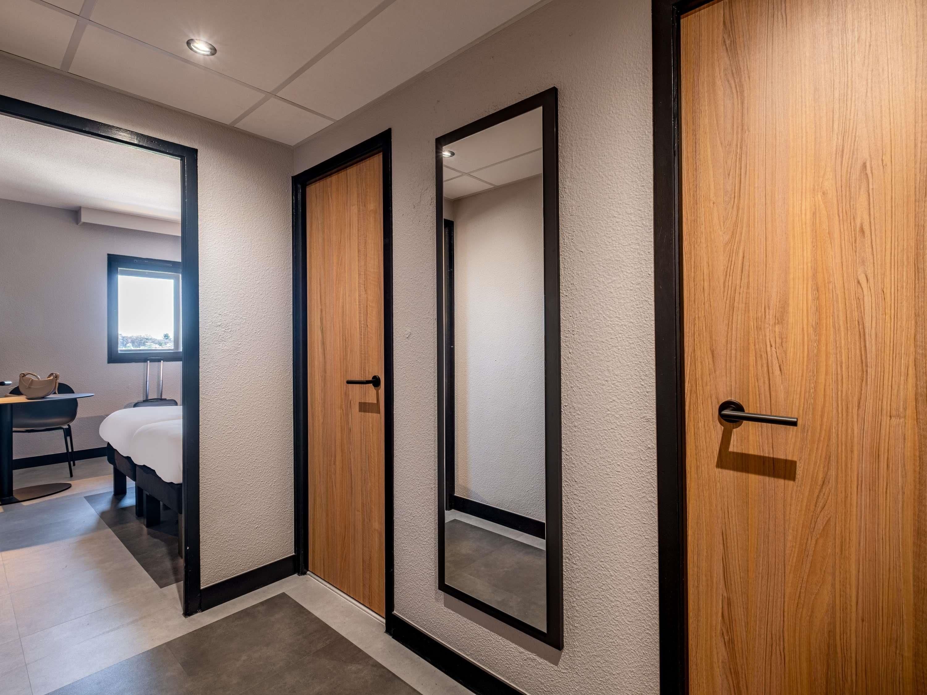 Un pasillo con dos puertas de madera y un espejo, que da a una habitación con cama y escritorio.