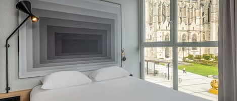 Habitación de hotel con una cama grande, una obra de arte moderna en la pared, una lámpara de pie y vistas a un edificio histórico a través de un ventanal amplio.