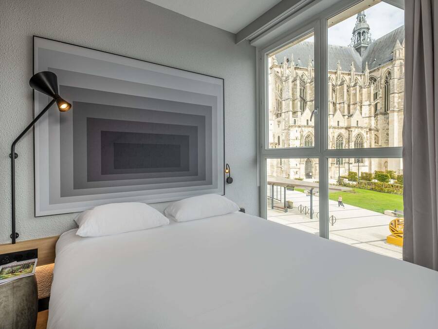 A hotel room with a large bed, a modern wall art piece, a floor lamp, and a view of a historic building through a large window.