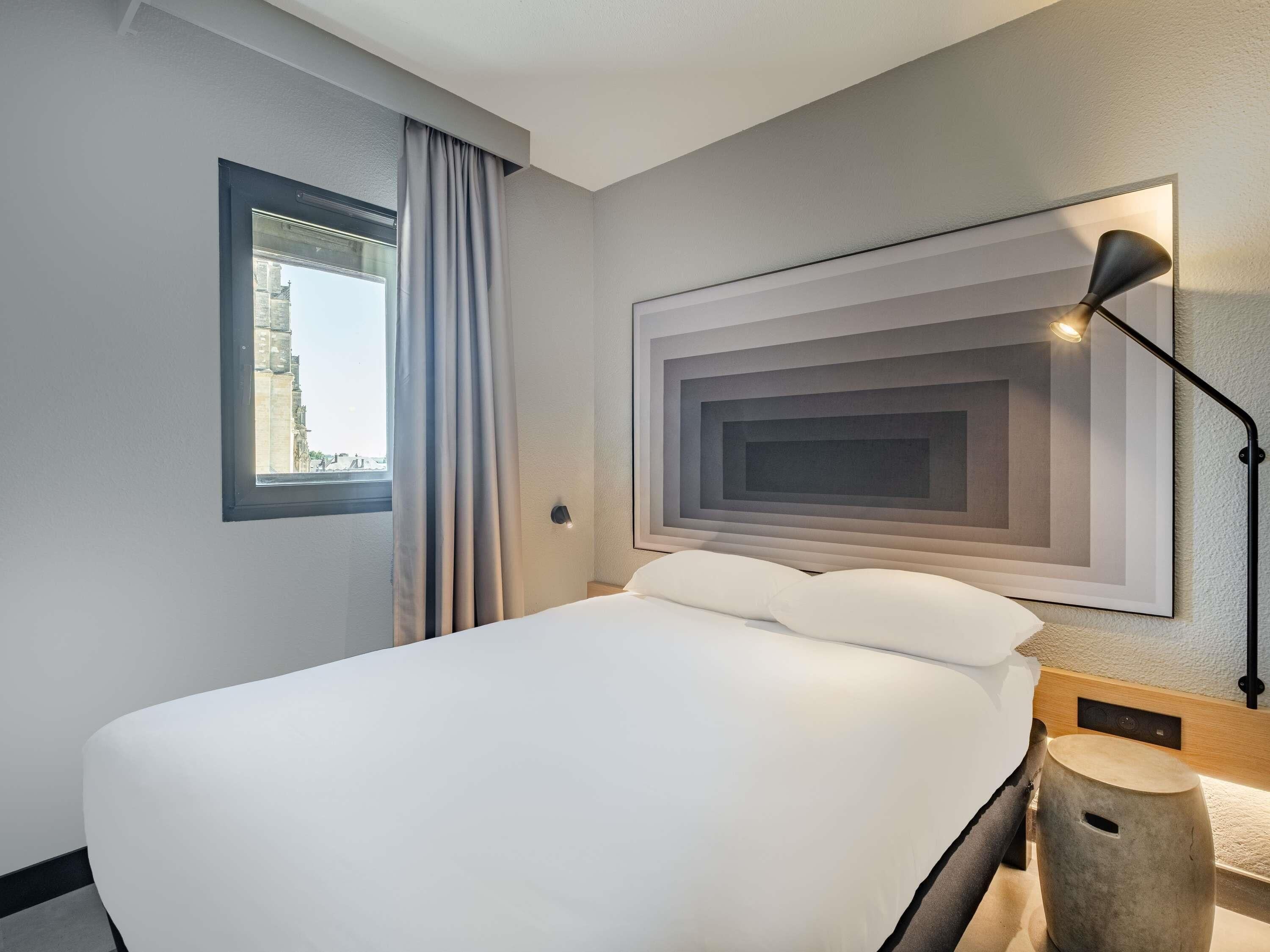A hotel room with a large bed, a window with curtains, a wall-mounted lamp, and a framed artwork.