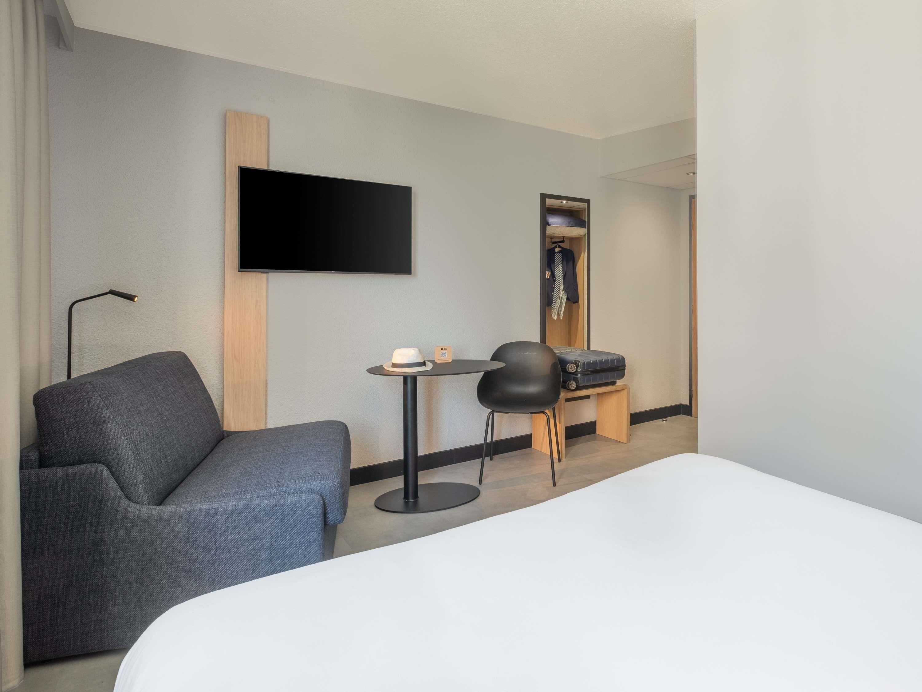 Una habitación de hotel con una cama, un sillón gris, una mesita con un sombrero y un teléfono, un televisor montado en la pared y un espejo con perchero.