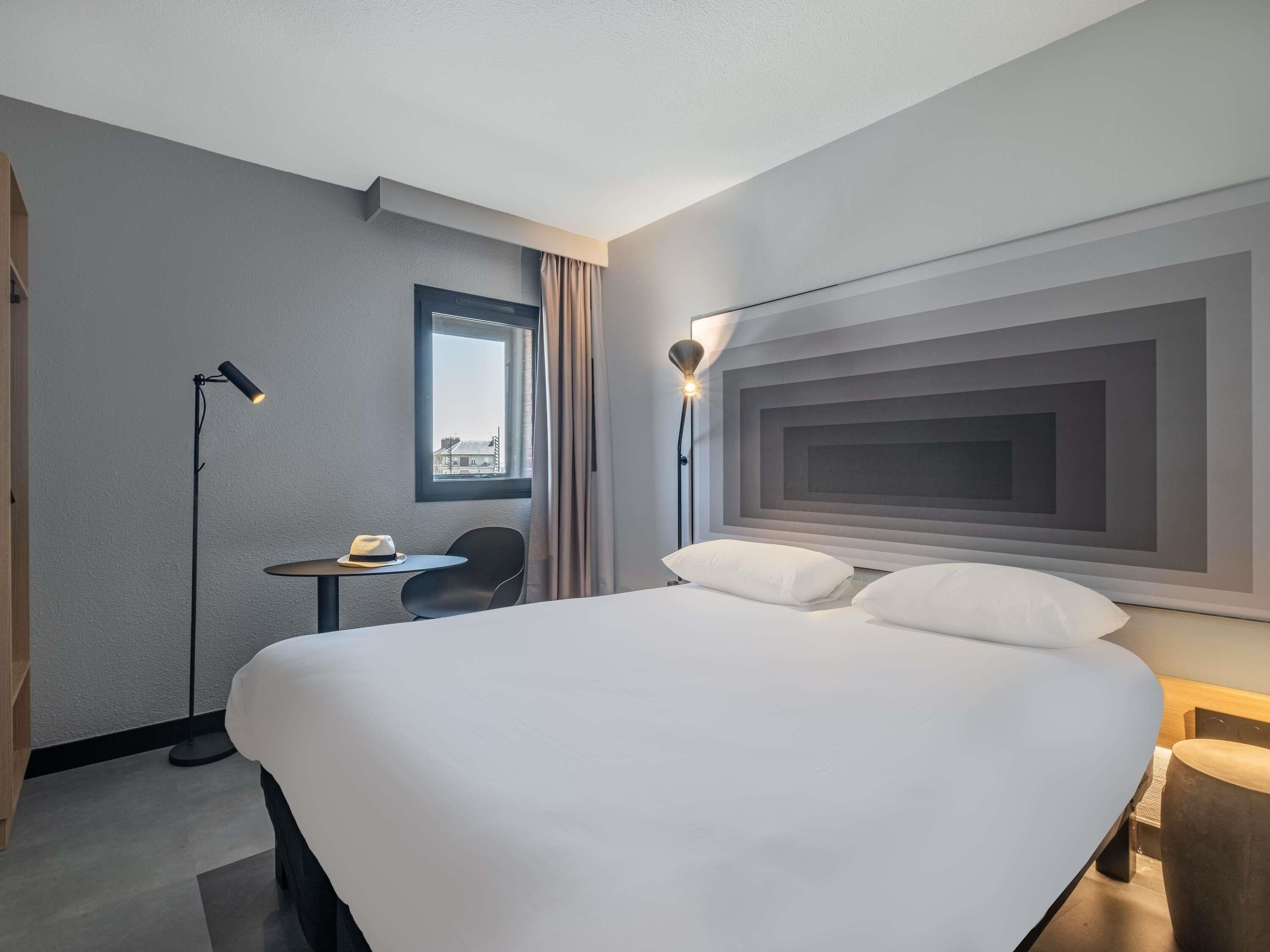 Habitación de hotel con una cama grande, un escritorio con una silla, una lámpara de pie y una ventana con cortinas.